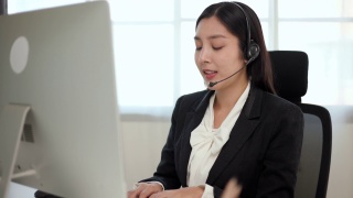 Smiley亚洲商务女接待员戴耳机视频会议电话，在电脑上通过网络摄像头在网上聊天，客户支持服务。视频素材模板下载