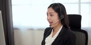 Smiley亚洲商务女接待员戴耳机视频会议电话，在电脑上通过网络摄像头在网上聊天，客户支持服务。