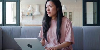 年轻沉思的亚裔女性坐在客厅的沙发上，在笔记本电脑上工作，打字，环顾四周，看起来心烦意乱，失望和沮丧