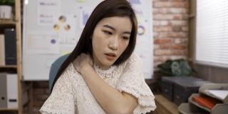 一名过度劳累的亚洲商务女性在办公室工作时，脸上带着痛苦的表情按摩着僵硬的肩膀肌肉。