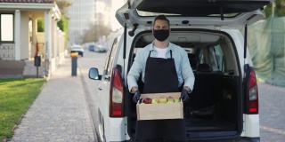 在户外的街道上，一个戴着面具、面带微笑的人拿着装着苹果的木箱，一个穿着围裙、戴着手套的快乐送货员站在箱子旁的肖像。网上购物配送，智能蔬菜快递服务，慢动作