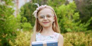 一个漂亮的7岁小女孩戴着眼镜，送了一个装在蓝色盒子里的带蝴蝶结的礼物。