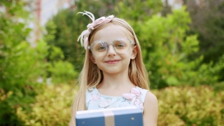 一个漂亮的7岁小女孩戴着眼镜，送了一个装在蓝色盒子里的带蝴蝶结的礼物。视频素材模板下载