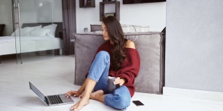 微笑的黑发女人在毛衣坐在地板上的咖啡和智能手机，而使用笔记本电脑