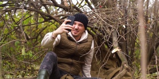 迷路的旅行者男性穿着暖和的衣服，坐在森林里，试图在阴冷的天气捕捉手机信号。