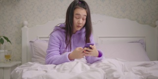 20多岁的亚洲女性晚上坐在床上用手机上网。穿着紫色睡衣的女人在床上用手机上网时打呵欠。