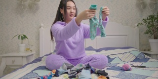 年轻的亚洲妇女折叠彩色袜子坐在床上。穿着紫色睡衣的女性通过整理衣服来整理和简化家庭。寻找袜子pair。