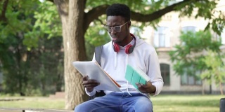 放大到压力过重的年轻学生坐在长凳上，手捧着平板电脑和文书。肖像的悲伤过度工作的非洲裔美国人叹息学习在大学院子。