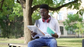 放大到压力过重的年轻学生坐在长凳上，手捧着平板电脑和文书。肖像的悲伤过度工作的非洲裔美国人叹息学习在大学院子。视频素材模板下载