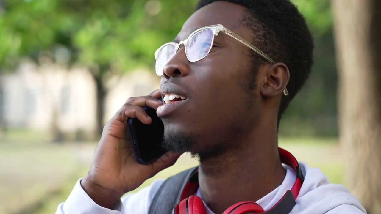 一幅心满意足的非裔美国人的头像，他戴着眼镜，坐在户外打电话。近距离的自信的聪明的男学生聊天在休息在大学校园。