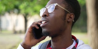 一幅心满意足的非裔美国人的头像，他戴着眼镜，坐在户外打电话。近距离的自信的聪明的男学生聊天在休息在大学校园。