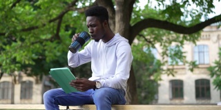 一名自信、专注的非洲裔美国人坐在大学院子的长凳上，喝着早茶，用平板电脑上网。集中的聪明英俊的学生在网上学习的肖像。