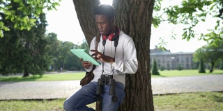 年轻专注的非洲裔美国学生倚在树干上用平板电脑上网。在阳光明媚的春天早晨，一名聪明的男子在户外检查作业。