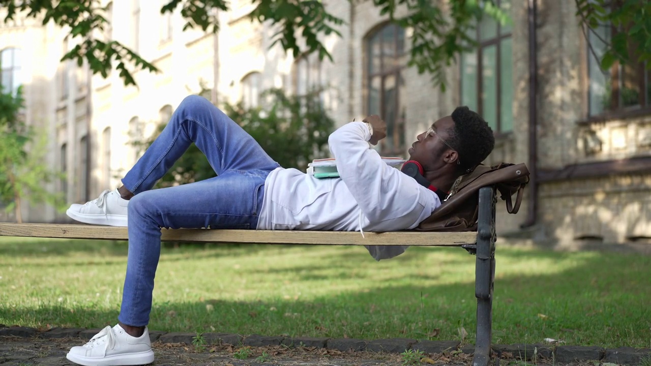 睡在户外长椅上的非洲裔美国学生疲惫不堪，醒来后急忙跑开，离开。年轻聪明疲惫的男人背着背包和文书奔向学习。教育和疲劳。