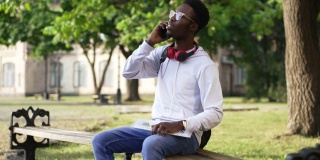 一名积极聪明的非洲裔美国学生，戴着眼镜，坐在长凳上讲电话，面带微笑。一幅英俊聪明的年轻人的肖像，在大学校园的户外。教育。