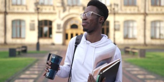 一个疲惫的非洲裔美国人站在大学校园里拿着书和空的热水瓶打呵欠。这是一幅在户外彻夜未眠、疲惫不堪的年轻学生的肖像。教育和生活方式。