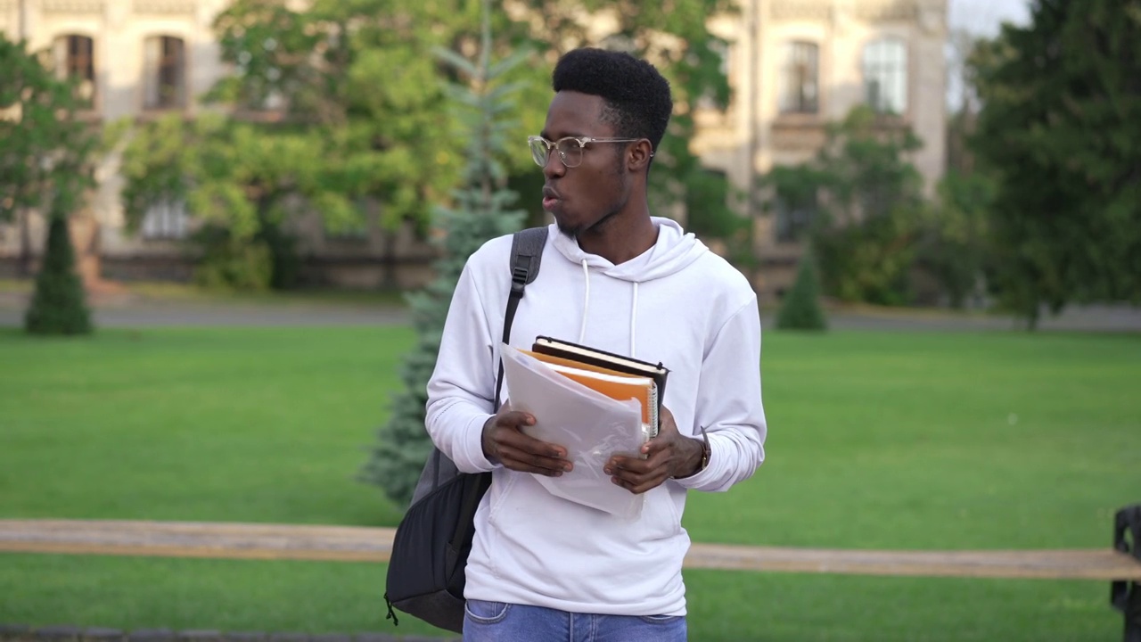 年轻英俊的非裔美国人，戴着眼镜，站在户外，和一群人一起做文书工作。自信聪明的男学生的肖像在大学校园的早晨。