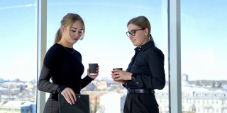 有吸引力的年轻女商务人员交流。穿着黑色衣服的女商人在办公室窗口的背景上喝饮料和谈项目。
