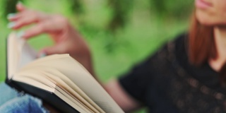 一个女人拿着一本书坐在公园的一棵树旁。一个穿着牛仔裤和t恤的女人在户外看书。