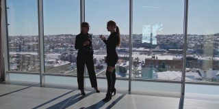 女性在工作中的沟通。两个年轻迷人的女人站在窗前讨论着什么。阳光明媚的办公室，大窗户。