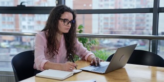 女商人坐在宽敞的办公室里，用笔记本电脑工作，做笔记。戴眼镜在电脑上查找信息的女人。