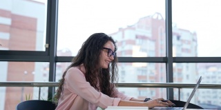 漂亮的女人在电脑前微笑着工作。女员工心情好，在办公室工作。工作场所在大灯光办公室和大窗户。