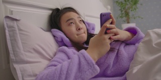 快乐的千禧一代亚洲妇女躺在床上用手机打字。年轻的成年女性穿着睡衣用手机上网。手持相机。