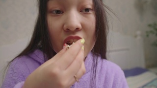 近距离的亚洲妇女吃爆米花，而看电影或电视节目。穿紫色睡衣的女士在吃爆米花。手持相机。视频素材模板下载