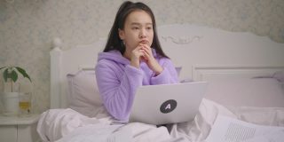 漂亮的亚洲女人坐在床上用笔记本电脑思考。辞职的女性有一个好主意，在笔记本电脑上打字。