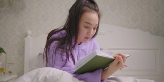 20多岁亚洲女性独自坐在床上看书。穿着紫色睡衣的女性喜欢坐在床上看书。
