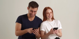 工作室肖像的年轻男子显示有趣的视频在智能手机上快乐的女朋友站在白色孤立的背景。沉迷于手机的情侣用手机看屏幕，慢动作。