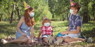宽镜头快乐的白人兄弟姐妹坐在春天夏天的公园野餐聊天。戴着派对帽和冠状病毒口罩的红发男孩女孩在阳光下户外庆祝生日。