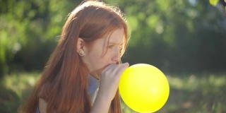 侧视图无忧无虑的少年红发女孩吹黄色气球坐在阳光在春天夏天的公园。轻松自信的高加索青年肖像与红色头发享受休闲户外。