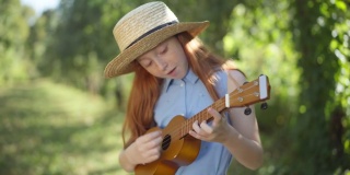 中镜头的肖像快乐熟练的高加索红发女孩唱歌玩尤克里里户外。阳光明媚的日子里，自信的青少年音乐家在春夏季的公园里享受业余爱好。人才和生活方式。