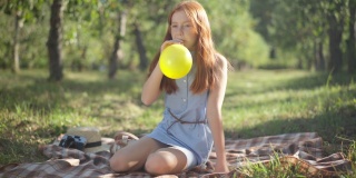 宽镜头肖像的红发美丽的少女吹气球坐在毯子在春天夏天的公园。自信轻松的高加索青少年长发享受周末休闲阳光。