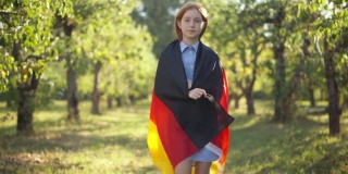 自信自豪的红发女孩包装在德国国旗走在阳光下看着相机。美丽的白人少年的肖像在春天夏天公园户外散步。多莉。