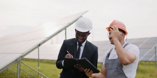 站在太阳能工厂中的技术员和检验员。戴着黄色安全帽和安全眼镜的印度工程师拿着数码平板电脑，与太阳能工厂的男性老板交谈。检查的能量