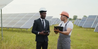 业主和工人在户外的太阳能电池间签署文件。穿着黑色西装和白色头盔的非裔美国人在夹板上签署文件，而夹板上则是穿着制服的印度人。光伏电池