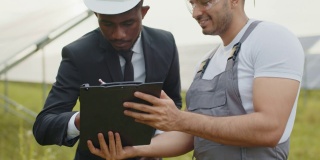 近距离拍摄西装革履的非洲人在夹着印度工程师的纸板上签署文件。两个多种族的人站在太阳能农场并达成协议。男子和技术人员在太阳能电池