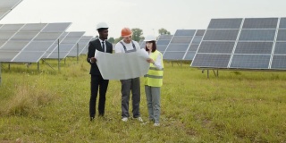 一群多民族的人戴着安全帽盯着太阳能农场，看着图纸。2名工程技术人员审核室外光伏建设方案。太阳能工厂工人