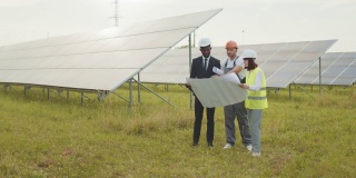 人们戴着安全帽站在户外的太阳能电池之间，开着工作会议。有能力的工人讨论生产替代能源的工厂。太阳能农场计划的工人