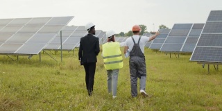 两个戴着白色头盔的检查人员和穿着制服的技术人员在太阳能电站上行走的背影。多种族工程师在户外检查光伏电池的工作。工人们在太阳能电站上行走
