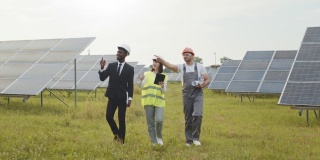 工人们在太阳能电站上行走。两名戴着白色头盔的检查人员和穿着制服的技术员在太阳能电站上行走。多种族工程师在户外检查光伏电池的工作
