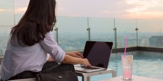夏日生活方式亚洲妇女放松和chill附近的豪华游泳池摩天大楼。一个亚洲女人在笔记本电脑上工作，模糊的背景摩天大楼，美丽的天空。