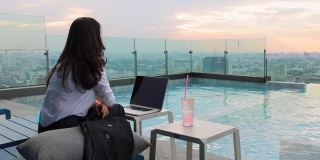 亚洲妇女在工作日缺乏精力。疲惫的女商人感到无聊，坐在摩天大楼的泳池边，拿着笔记本电脑和荔枝鸡尾酒。美丽的日落在背景。