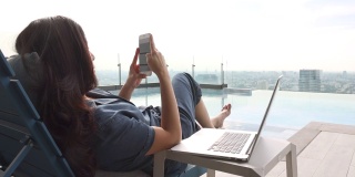 在明亮的阳光下，亚洲女人平静地放松在靠近游泳池的摩天大楼的椅子上，使用笔记本电脑，在应用程序智能手机和模糊背景城市上选择最喜欢的音乐。