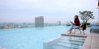 亚洲女性旅行者计划暑假和在平板电脑上搜索信息，而坐在摩天大楼的游泳池对美丽的风景背景与你的广告文本信息的拷贝空间。