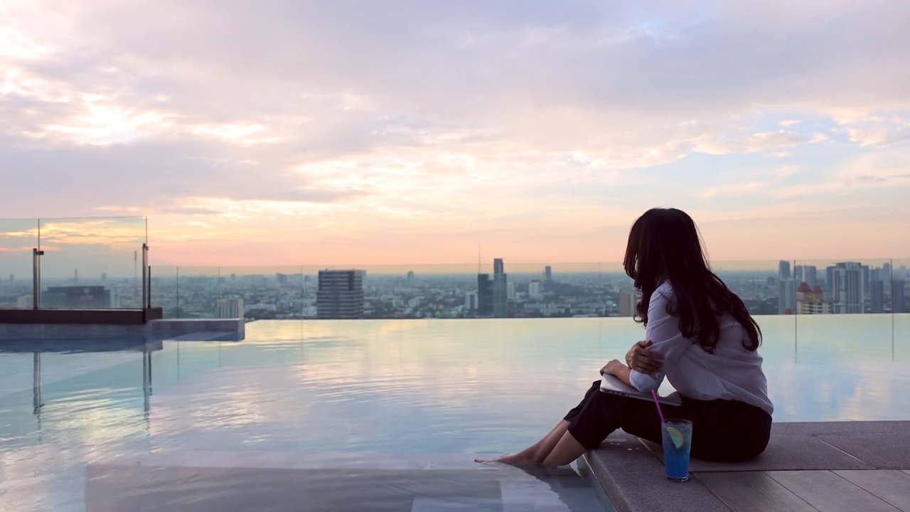 亚洲女商人坐在池边与她的脚在水里。女人一边工作，一边在笔记本电脑上打字，一边欣赏着城市摩天大楼里美丽的日落或日出天空。