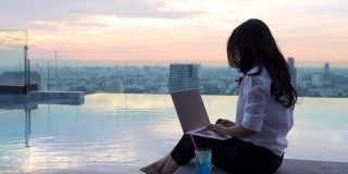 快乐的亚洲女人放松与笔记本电脑坐在边缘摇晃她的脚在游泳池看着远离梦想休息与城市的日落时间。
