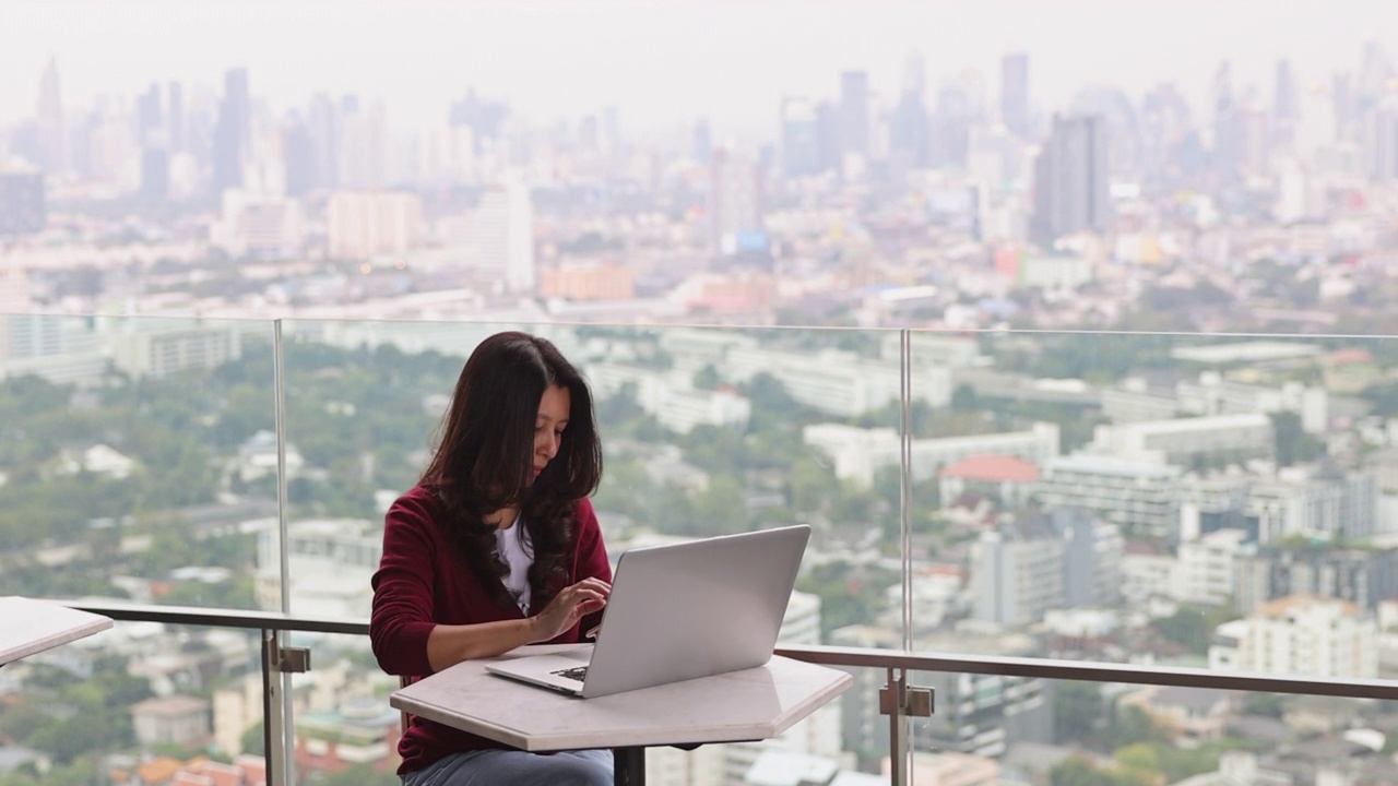 亚洲女性自由职业者坐在摩天大楼上用笔记本电脑工作，用智能手机在线订餐。忙碌，在假期努力工作。